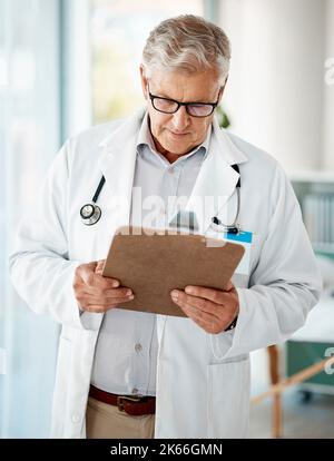 Handsome mature male caucasian doctor in white lab coat looking out at paperwork on a clipboard. One senior man holding medical or patient records Stock Photo
