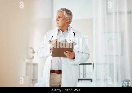 Pensive mature male caucasian doctor in white lab coat looking out window, thinking or pondering. One senior man holding a clipboard with patient Stock Photo
