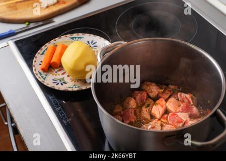 Pot over the fire of a ceramic hob cooking beef for a homemade stew, with potatoes and carrots prepared. Stock Photo