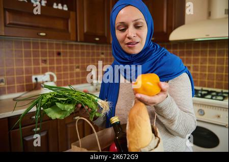 Happy young Muslim Arab housewife lays out purchases in her kitchen. Beautiful woman in good mood is sorting vegetables Stock Photo