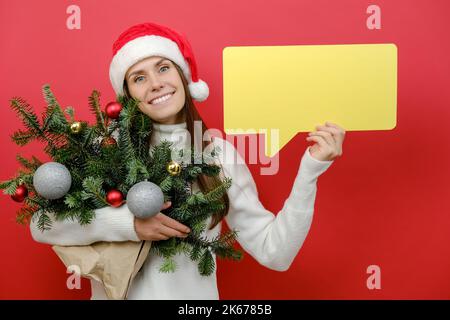 Portrait of happy young female in Christmas hat holding bouquet of spruce branches and yellow empty blank Say cloud, speech bubble posing on red backg Stock Photo