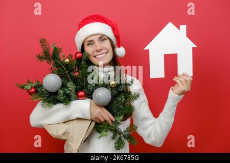 Smiling attractive young woman in Santa Claus hat holding bouquet of spruce branches and house model, posing over red studio background with copy spac Stock Photo