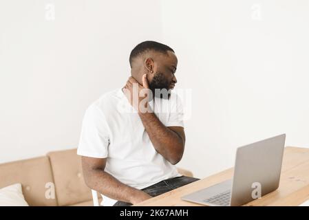 Portrait of a tense handsome black man with neck pain. Man working on business project using laptop suffering from neck pain. Stock Photo