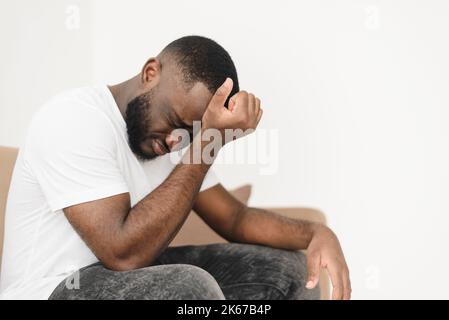 Stressed bearded african american man suffering from headache and sitting on sofa at home and rubbing his forehead, copy space. Upset man suffering fr Stock Photo