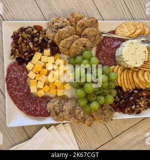A charcuterie platter of cheese, grapes, nuts, sausage, cookies, crackers and cheese spread on a white platter sitting on a table. Stock Photo