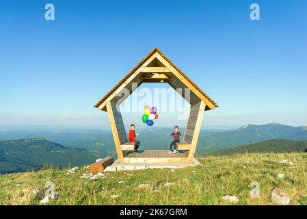 Girl and a boy are sitting in a wooden house on a mountain holding balloons. Concept of security, insurance and bright future Stock Photo