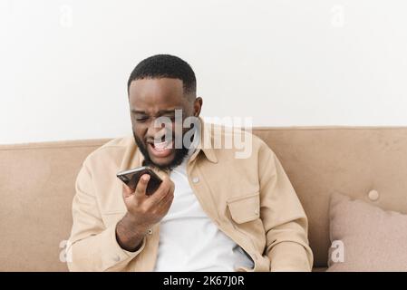 Annoyed african american man yelling at phone while sitting on sofa at home. Angry man looks at the phone while reading bad news. Business problems Stock Photo