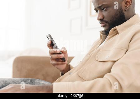 Annoyed african american man yelling at phone while sitting on sofa at home. Angry man looks at the phone while reading bad news. Business problems Stock Photo