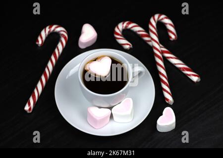 White cup of coffee or tea with marshmallow hearts and candy canes on dark wooden table. Hot drink with sweets for Christmas holidays Stock Photo
