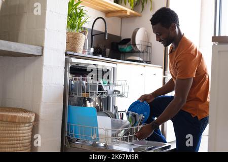 Happy african american man standing in kitchen and unloading dishwasher Stock Photo
