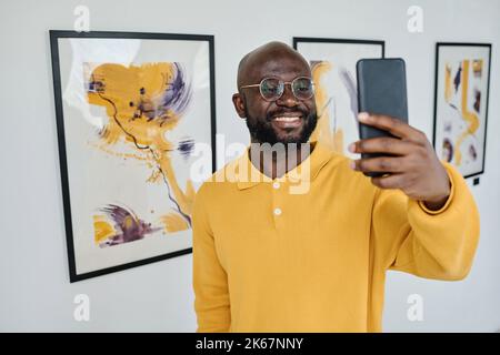 African young man in eyeglasses using his mobile phone for streaming standing at art gallery Stock Photo
