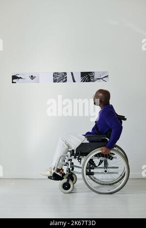 African young guy sitting in wheelchair and looking at modern photos on the wall at art gallery Stock Photo