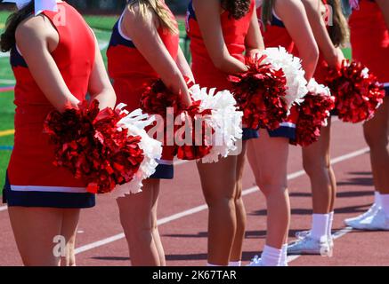 High school cheerleaders in red uniforms standing on the sideline holding their red and white pom poms behind them during a football game. Stock Photo