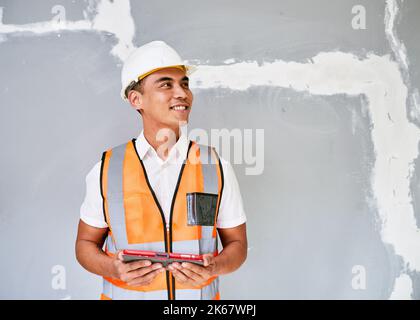 A young Asian construction worker looks to the right in front of cracked wall Stock Photo