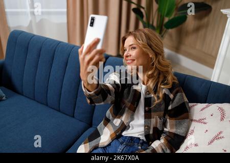 Pleasant middle-aged woman sitting on the sofa in the living room and taking a selfie on the cell phone Stock Photo