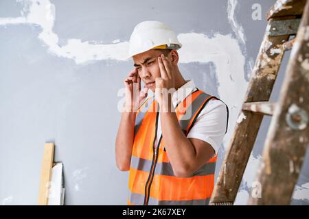 A young Asian construction worker rubs his temples in pain on site Stock Photo