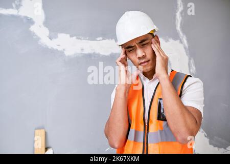 A young Asian construction worker rubs his temples in pain on site Stock Photo
