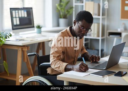 Portrait of black young man with disability working in office and using laptop, accessible workplace concept Stock Photo