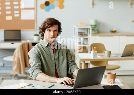 Portrait of long haired young man at office workplace using laptop and looking at camera with smile, IT professional, copy space Stock Photo