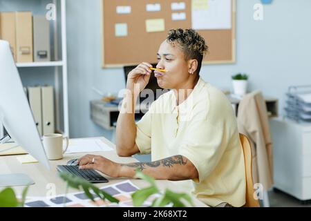 Portrait of tattooed black woman playing with pencil while using computer at office workplace Stock Photo