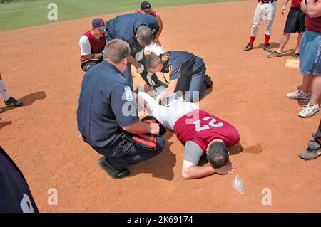 softball Player with broken leg is attended to by fire personal and wife as team members look on while laying on ball field during softball game Stock Photo