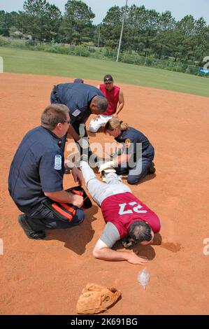 softball Player with broken leg is attended to by fire personal and wife as team members look on while laying on ball field during softball game Stock Photo