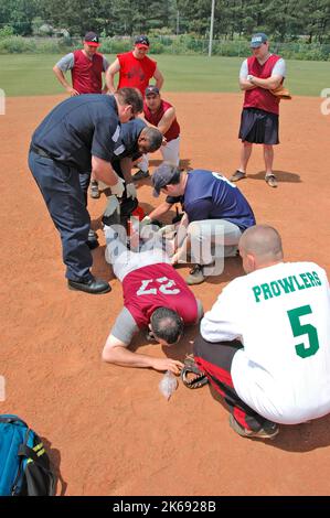 softball Player with broken leg is attended to by fire personal and wife as team members look on while laying on ball field during softball game Stock Photo