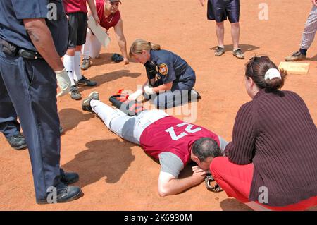 softball Player with broken leg is attended to by fire personal and wife as team members look on while laying on ball field during softball game Stock Photo