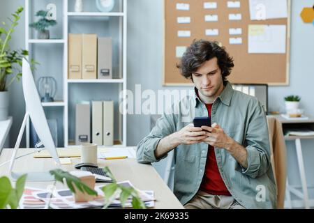 Portrait of Caucasian young man using smartphone at workplace and scrolling social media during break Stock Photo