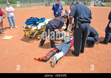 softball Player with broken leg is attended to by fire personal and wife as team members look on while laying on ball field during softball game Stock Photo