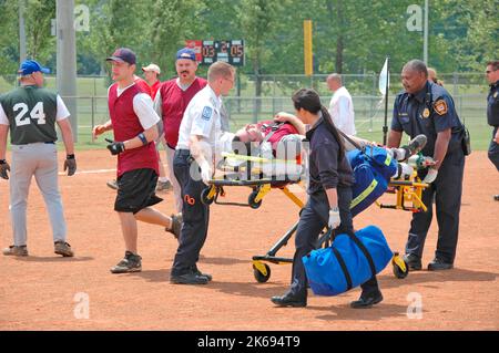 softball Player with broken leg is attended to by fire personal and wife as team members look on while laying on ball field during softball game Stock Photo