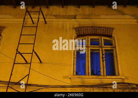 Window of old house viewed from outside with wind chimes and metal ladder on wall, lit by blue light from inside, in Bucharest Stock Photo