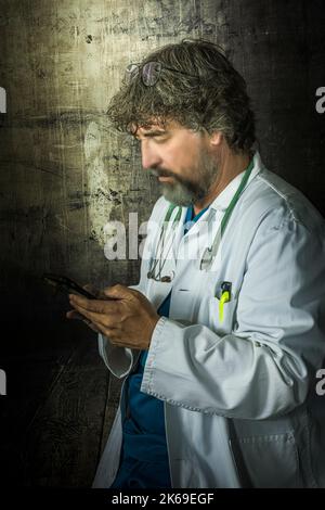bearded mature doctor dressed in a blue surgical suit and a white coat looking at the mobile phone distractedly thinking about answering healthcare me Stock Photo