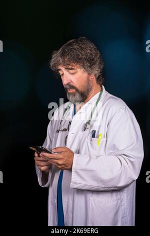 bearded mature doctor dressed in a blue surgical suit and a white coat looking at the mobile phone distractedly thinking about answering healthcare me Stock Photo