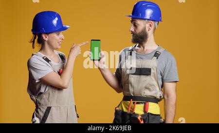 Portrait of building contractors pointing at smartphone with greenscreen display in studio, working on construction. Holding isolated mockup template with blank chroma key copyspace. Stock Photo