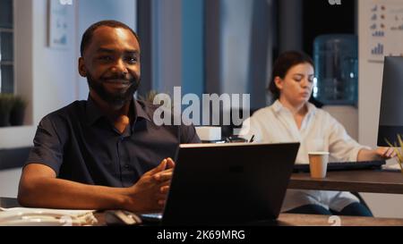 Portrait of happy successful manager sitting at desk at night, working on corporate presentation in office. Planning research data to create professional executive report with financial web app. Stock Photo