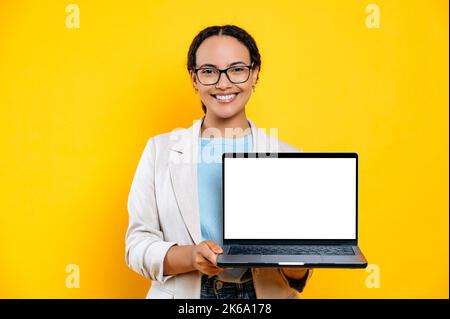 Gorgeous successful confident young brunette latino or brazilian woman, hold laptop computer with blank mock-up copy space screen, stand on isolated orange background, looks at camera, smiles friendly Stock Photo