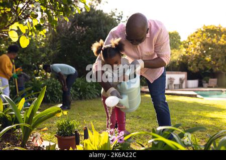 Happy senior african american man and his granddaughter watering plants in sunny garden Stock Photo