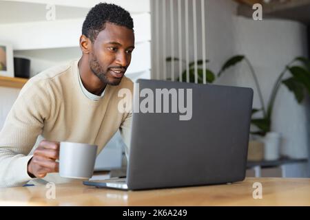 Happy african american man leaning on countertop in kitchen, using laptop Stock Photo