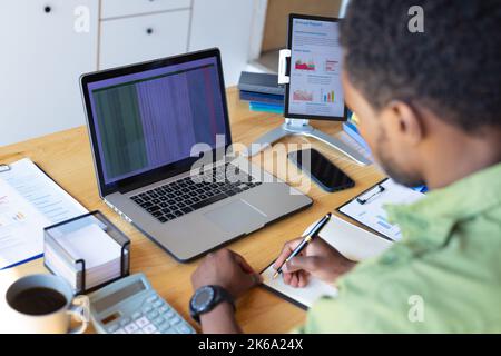 Happy african american man standing at table in kitchen, using laptop and taking notes Stock Photo