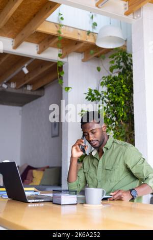 Happy african american man standing at table in kitchen, using laptop and talking on smartphone Stock Photo