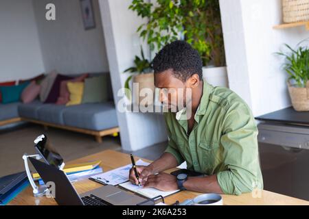 Happy african american man standing at table in kitchen, using laptop Stock Photo