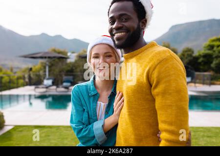 Portrait of happy diverse couple in santa hats to celebrate christmas smiling outdoors Stock Photo
