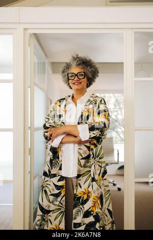 Senior businesswoman smiling at the camera while standing in an office with her arms crossed. Happy businesswoman working in a creative workplace. Stock Photo