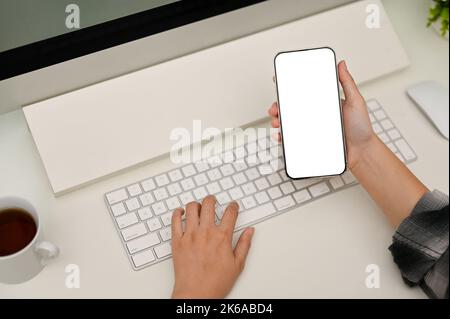Close-up image, Female hand holding a smartphone white screen mockup while typing on computer keyboard at her minimal white office desk. Stock Photo