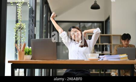 Overworked and tired young Asian businesswoman or female office worker sitting at her office desk, stretching her arms, suffering from shoulders pain. Stock Photo