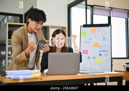 Happy and overjoyed Asian business colleagues triumph, looking at laptop screen, celebrating their success together, project winning, good job results Stock Photo