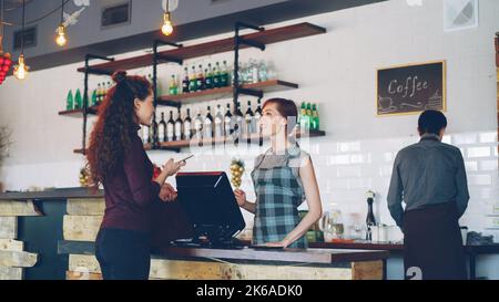 Young people are buying coffee-to-go in nice local cafe and paying with smartphone while friendly workers are greeting customers, talking and selling drinks. Stock Photo