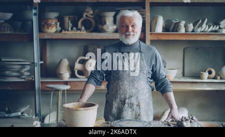 Portrait of senior silver-haired potter in muddy apron standing alone at table in workshop and looking at camera. Shelves with handmade vases and pots in background. Stock Photo
