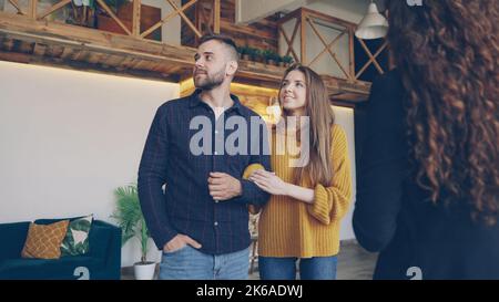 Confident housing agent is showing spacious modern house with beautiful interior to happy young couple wife and husband, people are smiling and talking looking around. Stock Photo